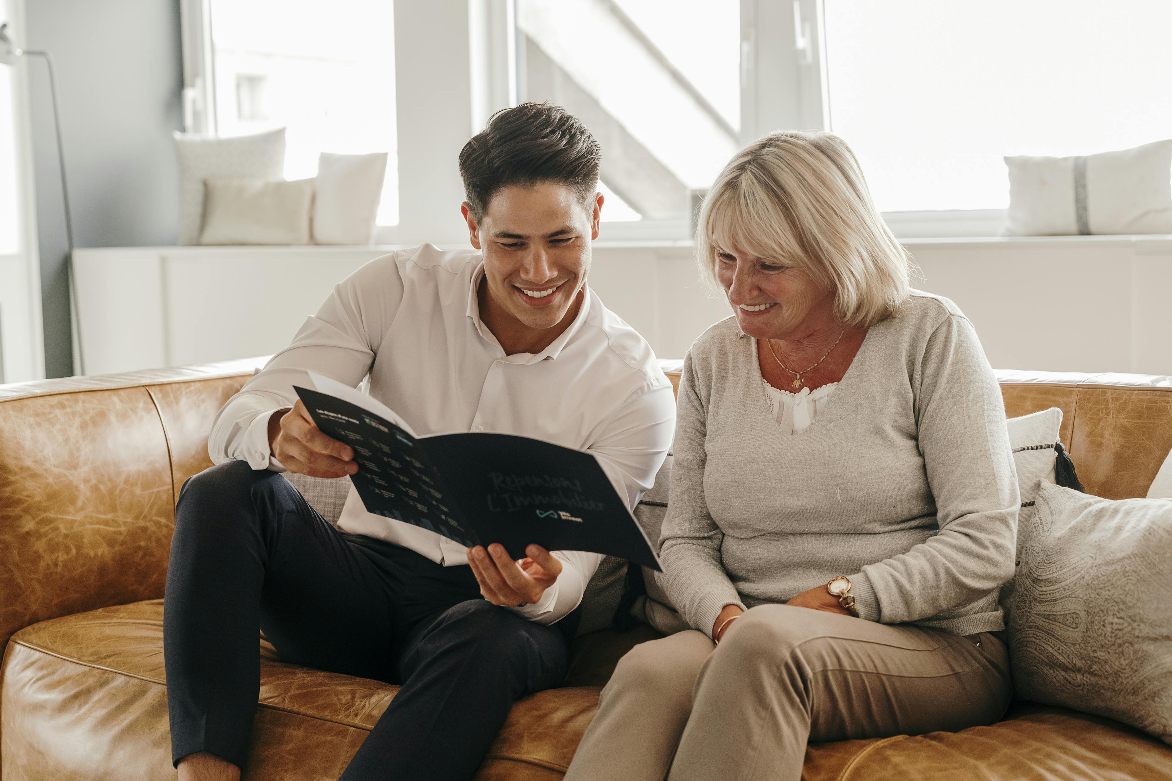 Assis sur un canapé en cuir marron, un agent immobilier We Invest explique à une cliente les différentes étapes d'une vente immobilière avec une brochure en support.